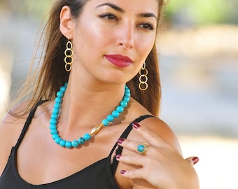 May include: A woman wearing a black sleeveless top with a turquoise beaded necklace, gold hoop earrings, a turquoise ring, and a gold bracelet with a turquoise stone.
