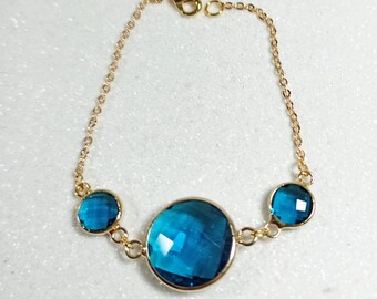 May include: A gold chain bracelet with three blue gemstone charms. The largest charm is round and the two smaller charms are oval.