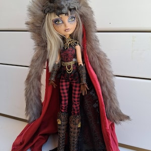 Puede incluir: Una muñeca con cabello rubio y maquillaje llamativo, que lleva un tocado de lobo y un atuendo a cuadros rojos. La muñeca tiene una larga capa con ribete de piel y botas marrones. La muñeca está sobre un soporte negro.