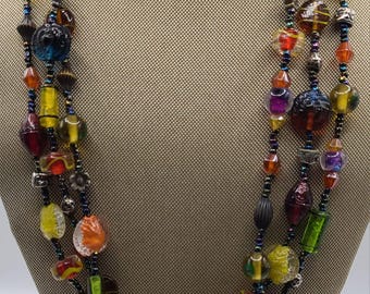 May include: Multi-strand necklace with colorful glass beads in shades of red, orange, yellow, green, and blue. The beads are of various shapes and sizes, including round, oval, and cylindrical. The necklace is strung on a black cord.