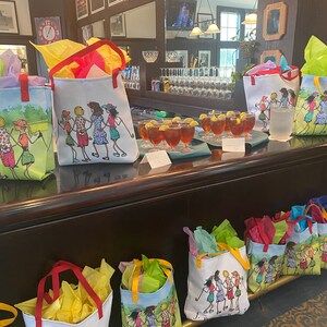 May include: A table with several colorful tote bags with a design of women playing golf. The bags are filled with tissue paper and are ready to be given as gifts. There are also glasses of iced tea on the table.