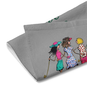 May include: Gray golf towel featuring a colorful illustration of three women in golf attire. The women are holding golf clubs and wearing hats and dresses. The towel is made of a soft, absorbent material.