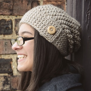 May include: A woman wearing a beige crochet beanie with a wooden button. The beanie has a slouchy fit and is crocheted in a textured stitch. The text "CROCHET PATTERN" and "littlemonkeyscrochet" is visible in the image.