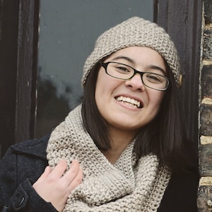 May include: A woman wearing a beige crochet infinity scarf and a beige crochet beanie. The scarf and beanie are both made with a simple stitch pattern. The woman is smiling and looking at the camera. The image is captioned "CROCHET PATTERN littlemonkeyscrochet"