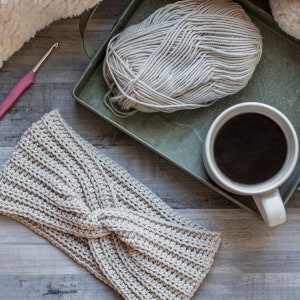 May include: A handmade, light beige crochet headband with a twisted knot design. A ball of yarn, a crochet hook, and a mug of coffee are also visible, suggesting a crafting theme. The headband is made of a ribbed stitch.