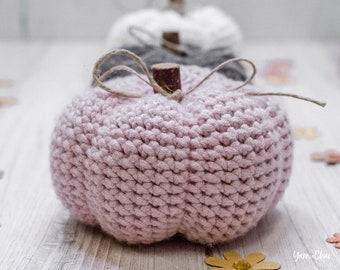 May include: A pink knitted pumpkin with a brown wooden stem and twine accents. The pumpkin is sitting on a white wooden surface with gold glitter scattered around it.