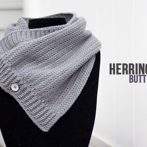May include: A gray crocheted cowl with a herringbone stitch pattern and two buttons. The cowl is displayed on a black mannequin.