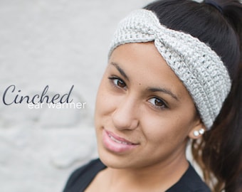 May include: A close-up of a woman wearing a gray crocheted ear warmer headband. The headband is twisted and has a simple design. The text "Cinched ear warmer" is visible in the image.