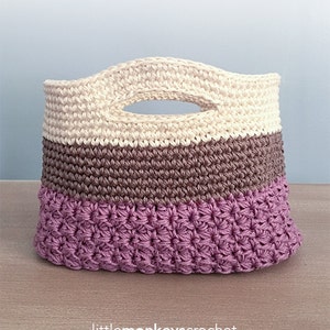 Pode incluir: Uma bolsa de presente de crochê com um design listrado creme, marrom e roxo. A bolsa tem uma alça e o texto "Huntley Gift Bag" está escrito acima da bolsa.