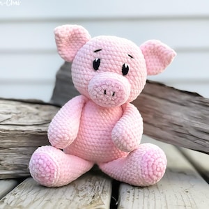 May include: A pink crocheted pig soft toy with black eyes and a pink snout. The pig is sitting on a wooden surface.