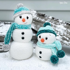 May include: Two crocheted snowmen with teal hats and scarves. The larger snowman has a white body with two black buttons and a carrot nose. The smaller snowman has a white body with two black buttons and an orange carrot nose.