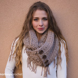 May include: A brown crocheted infinity scarf with fringe and two large buttons. The scarf is worn by a person with long brown hair.