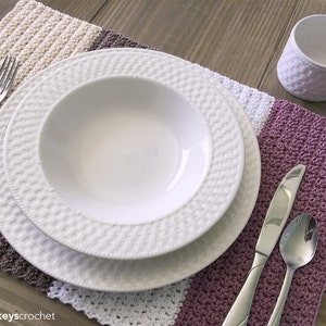 Puede incluir: Un juego de manteles individuales de ganchillo en beige, marrón, blanco y morado con un plato blanco, un tazón blanco, un tenedor de plata, un cuchillo de plata, una cuchara de plata y una taza de cerámica blanca.