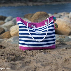 May include: A striped crochet beach bag with pink trim. The bag is sitting on a sandy beach with rocks in the background.