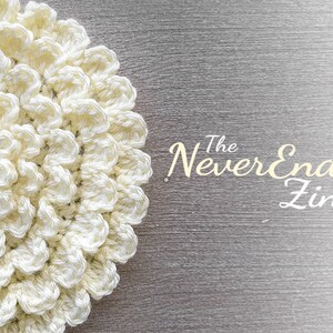 May include: A close-up of a crocheted white flower with a textured surface. The flower is on a gray background with the text "The NeverEnding Zinnia".