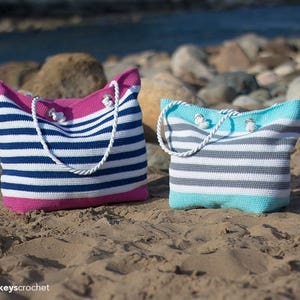 May include: Two striped crochet tote bags on a sandy beach. One bag is pink and white with blue stripes, the other is turquoise and white with gray stripes.