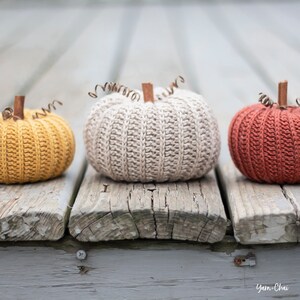 Op de afbeelding: Drie gebreide pompoenen in verschillende kleuren: geel, beige en oranje. De pompoenen liggen op een houten oppervlak.