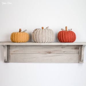 Op de afbeelding: Drie gehaakte pompoenen in verschillende kleuren, geel, beige en rood, worden op een houten plank getoond.