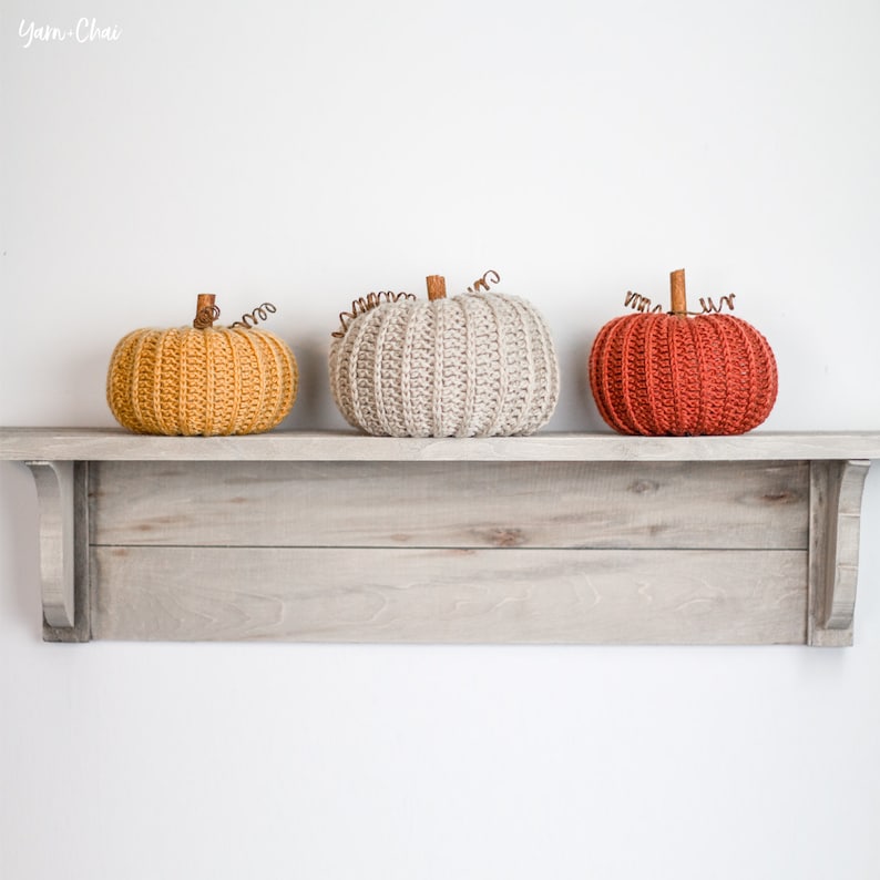 Op de afbeelding: Drie gehaakte pompoenen in verschillende kleuren, geel, beige en rood, worden op een houten plank getoond.