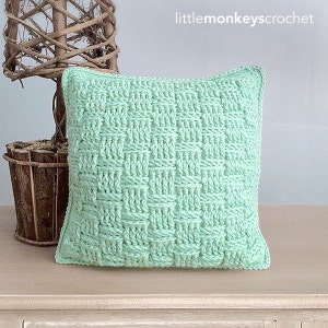 May include: A light green crocheted pillow with a woven pattern. The pillow is sitting on a light wood surface.