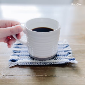 May include: A white ceramic mug filled with coffee sits on a blue and white woven coaster. The mug has a textured design and a gray base.