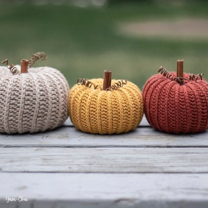 Op de afbeelding: Drie gehaakte pompoenen in beige, geel en oranje. De pompoenen liggen op een verweerd houten oppervlak.
