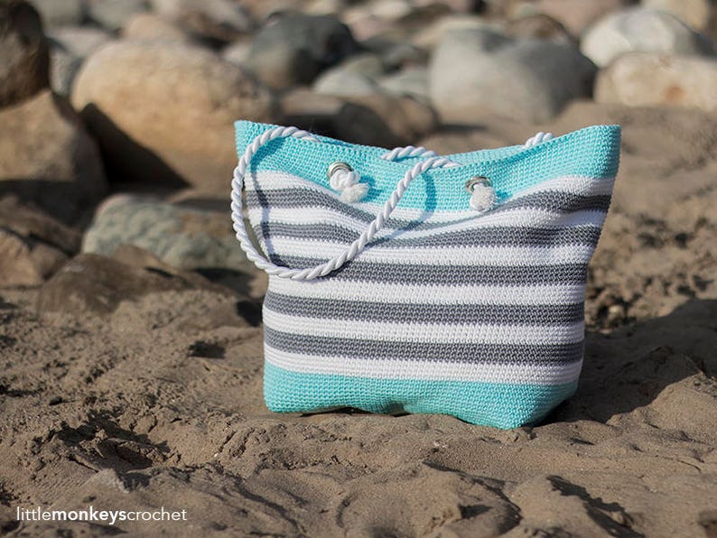 May include: A striped crochet tote bag with white and gray stripes and a light blue bottom. The bag has white rope handles and is sitting on a sandy beach.