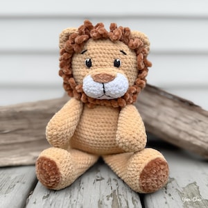 May include: A handmade crochet lion plush toy. The lion is primarily tan with a brown mane and accents. It has a white muzzle, black eyes, and a friendly expression. The plush is sitting on a weathered wooden surface.