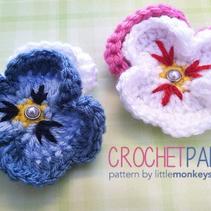 Könnte beinhalten: Zwei gehäkelte Stiefmütterchen-Blumen, eine blaue und eine weiße mit einem rosa Zentrum. Die Blumen haben eine Perle in der Mitte und sind mit schwarzen und roten Stickereien verziert. Der Text "CROCHETPANSY pattern by littlemonkeyscrochet" befindet sich unter den Blumen.