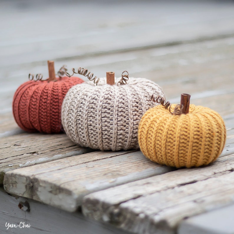 Op de afbeelding: Drie gehaakte pompoenen in verschillende kleuren: oranje, beige en geel. De pompoenen liggen op een houten oppervlak.