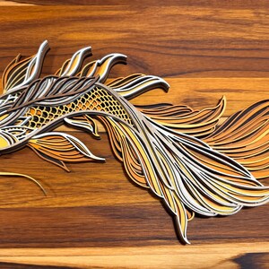 May include: A layered wooden art piece depicting a koi fish. The fish features intricate details with shades of yellow, orange, and white, set against a dark wood grain background. The artwork showcases a detailed, layered design.