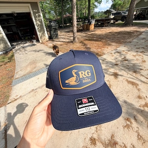Puede incluir: Gorra de camionero azul marino con un parche rectangular con las letras "RG" y un gráfico de pato, con el año "2021". La gorra tiene una visera curva y una parte trasera de malla. La etiqueta dice "Richardson 112 Trucker".
