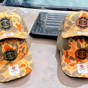 May include: Four camouflage trucker hats with orange, yellow, and brown patterns. Each hat features a circular patch with "ES" and a gear design. The hats have a mesh back and a curved brim. The label reads "112PFP".