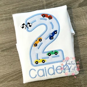 Puede incluir: Un pelele blanco con un n&uacute;mero dos azul bordado en &eacute;l. El n&uacute;mero dos est&aacute; bordeado de azul y tiene un patr&oacute;n a rayas azul y blanco en su interior. Hay cinco coches peque&ntilde;os bordados en el n&uacute;mero dos, cada uno de un color diferente. El nombre "Caiden" est&aacute; bordado debajo del n&uacute;mero dos. El texto "katie did it crafts" est&aacute; bordado en la esquina inferior derecha.