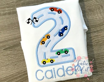 May include: A white onesie with a blue number two embroidered on it. The number two is outlined in blue and has a blue and white striped pattern inside. There are five small cars embroidered on the number two, each a different color. The name "Caiden" is embroidered below the number two. The text "katie did it crafts" is embroidered in the bottom right corner.