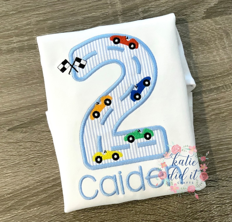 Puede incluir: Un pelele blanco con un n&uacute;mero dos azul bordado en &eacute;l. El n&uacute;mero dos est&aacute; bordeado de azul y tiene un patr&oacute;n a rayas azul y blanco en su interior. Hay cinco coches peque&ntilde;os bordados en el n&uacute;mero dos, cada uno de un color diferente. El nombre "Caiden" est&aacute; bordado debajo del n&uacute;mero dos. El texto "katie did it crafts" est&aacute; bordado en la esquina inferior derecha.