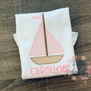 May include: A white onesie with a pink and brown sailboat design and the name "Caroline" embroidered in pink.