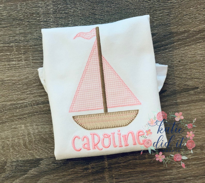 May include: A white onesie with a pink and brown sailboat design and the name "Caroline" embroidered in pink.