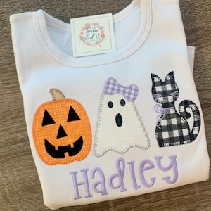 May include: White t-shirt with an orange pumpkin, a white ghost with a purple bow, and a black and white checkered cat. The shirt has the name "Hadley" embroidered in purple.