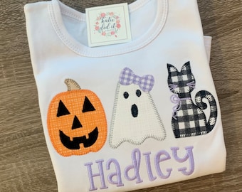 May include: White t-shirt with an orange pumpkin, a white ghost with a purple bow, and a black and white checkered cat. The shirt has the name "Hadley" embroidered in purple.
