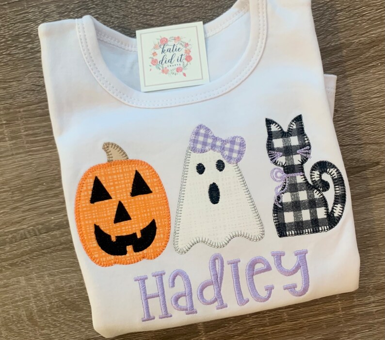 May include: White t-shirt with an orange pumpkin, a white ghost with a purple bow, and a black and white checkered cat. The shirt has the name "Hadley" embroidered in purple.