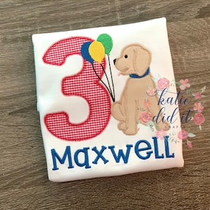 May include: A white t-shirt with a red and white gingham number "3" and a brown dog holding balloons. The text "Maxwell" is embroidered in blue below the number.
