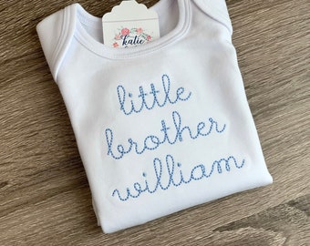 May include: White baby onesie with blue embroidered text that reads "little brother william".