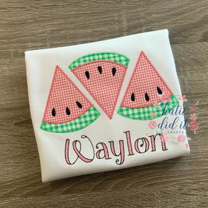 May include: White t-shirt featuring three watermelon slice appliqu&eacute;s. The slices have red and white gingham patterns, black seed details, and green gingham rind. The name "Waylon" is embroidered in pink script.