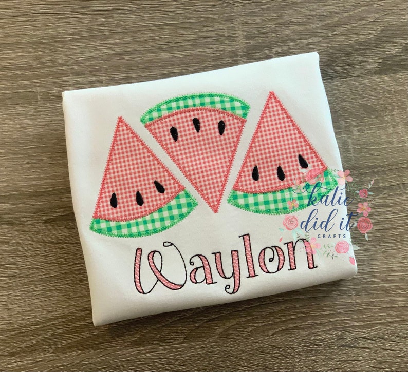 May include: White t-shirt featuring three watermelon slice appliqu&eacute;s. The slices have red and white gingham patterns, black seed details, and green gingham rind. The name "Waylon" is embroidered in pink script.