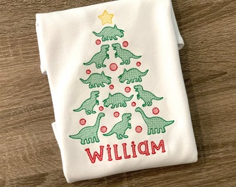 May include: White t-shirt with an embroidered green dinosaur Christmas tree with red ornaments and a yellow star on top. The name "William" is embroidered in red at the bottom of the tree.