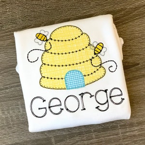 K&ouml;nnte beinhalten: Wei&szlig;er Baby-Strampler mit einem gelben und schwarzen Bienenstock und zwei Bienen, die auf der Vorderseite gestickt sind. Der Name "George" ist unter dem Bienenstock gestickt.