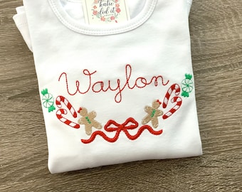 May include: White Christmas-themed t-shirt with the name "Waylon" embroidered in red thread. The design features gingerbread men, candy canes, and bows.