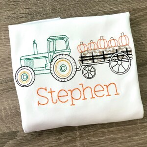 May include: White t-shirt with an embroidered design of a green tractor pulling a wagon filled with pumpkins. The name "Stephen" is embroidered in orange below the tractor.