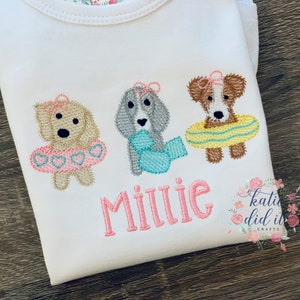 May include: White onesie with embroidered design of three dogs wearing floaties and bows. The name "Millie" is embroidered in pink. The design includes a light brown, gray, and brown dog. The onesie has a floral pattern on the neckline.